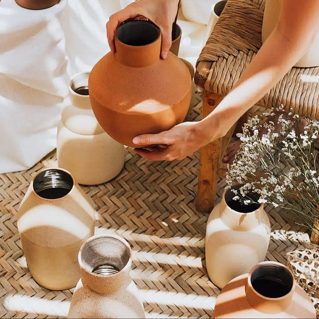 A person holds a terracotta-colored ceramic vase with a short neck and rounded body, surrounded by other matte ceramic vases in warm neutral tones on a woven mat.